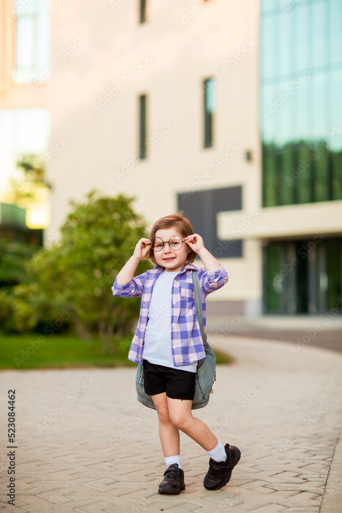 a little schoolgirl, standing in glasses,near the school building,with a backpack