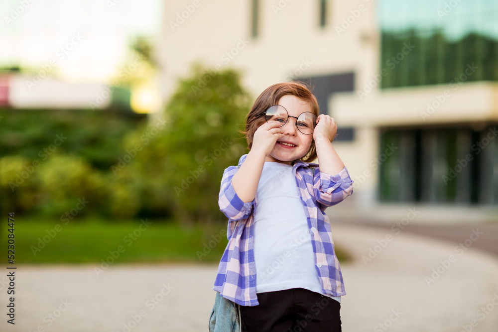 Fototapeta premium a little schoolgirl, standing in glasses,near the school building,with a backpack