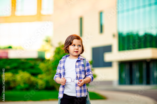 a little schoolgirl, standing in glasses,near the school building,with a backpack