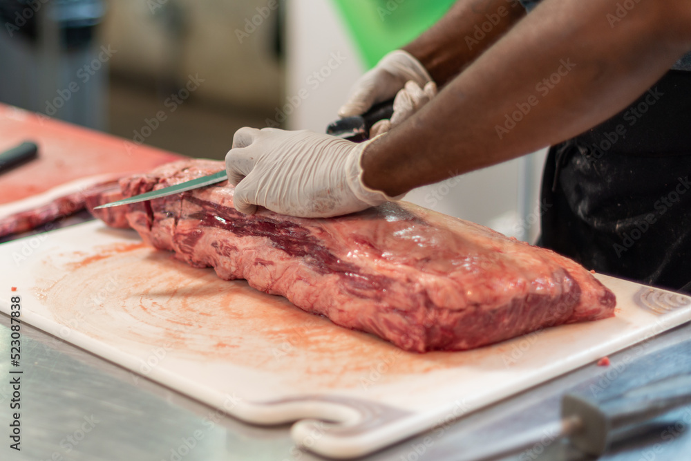 A close up of a large prime rib roast is sitting on a brown plastic cutting board. The meat has some marbling and multiple ribs. The chef is using a long knife to cut steaks from the fresh meat.