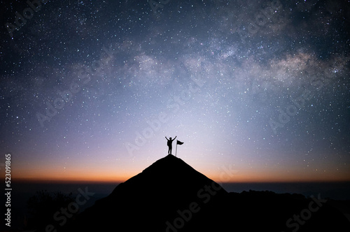 Silhouette of businessman standing on top of the mountain over night sky with star, Milky Way and sunlight with flag. He raised both arms, showing joy and success.