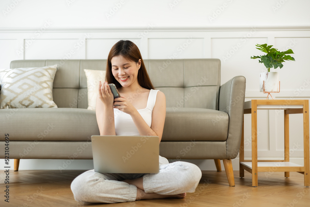 Freelance asian woman in glasses with mobile phone typing at laptop and working from home office. Happy girl sitting on couch in living room. Freelance asian woman in glasses with mobile phone typing at laptop and working from home office. Happy girl sitting on couch in living room.