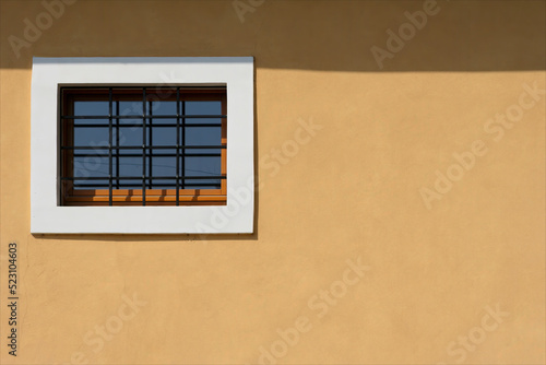 Particolare di facciata di edificio nuovo con muro giallo e una finestra sbarrata e con cornice di marmo bianco. Casa restaurata recentemente.  photo