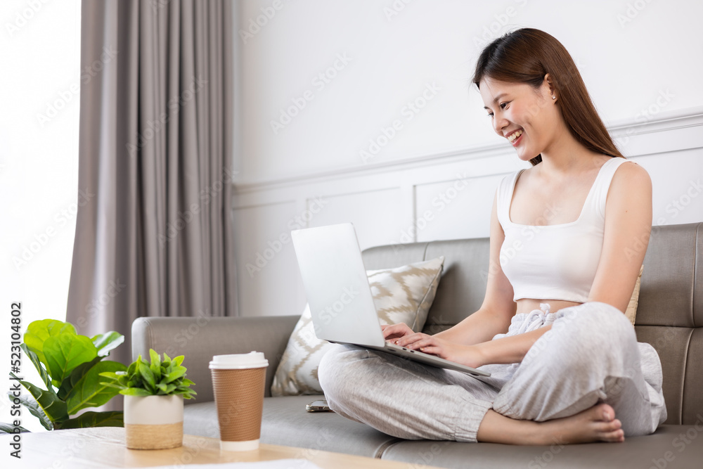Freelance asian woman in glasses with mobile phone typing at laptop and working from home office. Happy girl sitting on couch in living room. Freelance asian woman in glasses with mobile phone typing at laptop and working from home office. Happy girl sitting on couch in living room.