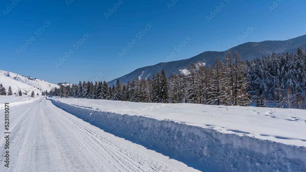 The highway, covered with trampled snow, goes into the distance. High snowdrifts on the roadsides. Coniferous forest and mountain range against the blue sky. Altai The highway, covered with trampled snow, goes into the distance. High snowdrifts on the roadsides. Coniferous forest and mountain range against the blue sky. Altai