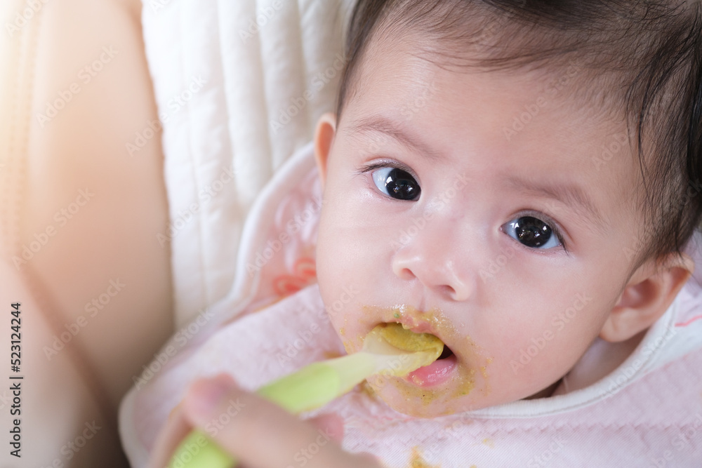 Cute baby girl looking at camara and eating food. Her mother giving healthy food by spoon in the morning at home.