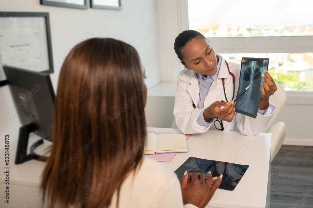 Confident doctor showing x-ray picture of lungs and explaining diagnosis to patient. Young African American pulmonologist consulting woman in clinic. Medical consultation, pulmonology concept Confident doctor showing x-ray picture of lungs and explaining diagnosis to patient. Young African American pulmonologist consulting woman in clinic. Medical consultation, pulmonology concept