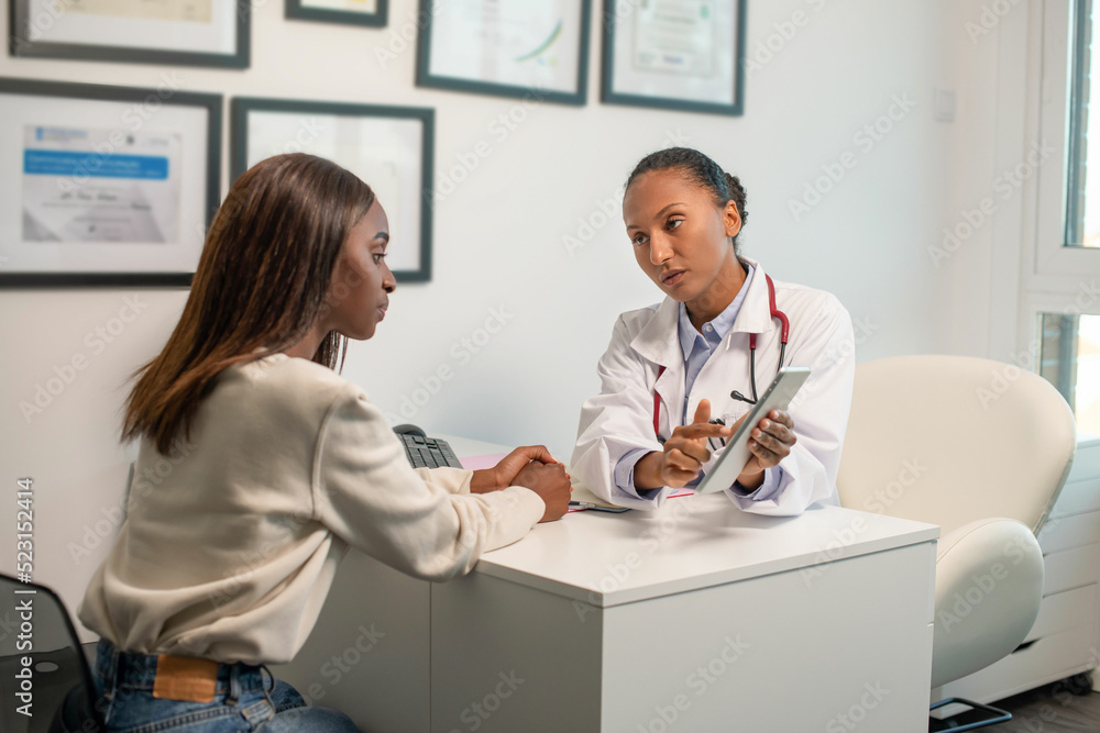 Obraz premium Confident doctor consulting female patient with digital tablet. Young African American woman sitting at doctors office and looking at her diagnosis on touchpad. Medical consultation concept