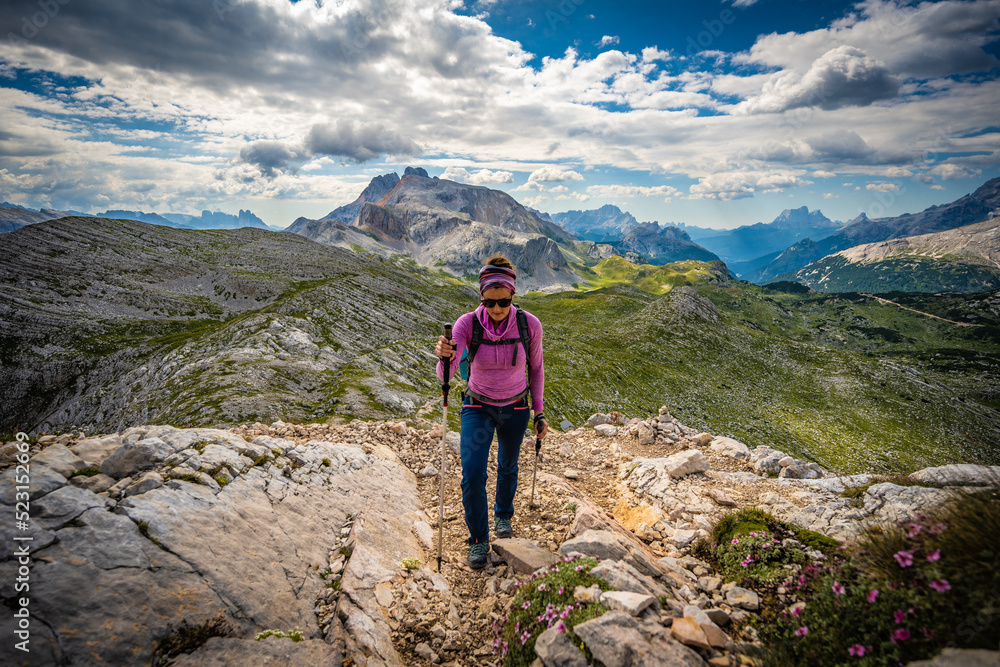 Young woman is hiking on beautiful trail with flowers from Baires Lake to Seekofel in the Dolomite mountains in the morning. Seekofel, Dolomites, South Tirol, Italy, Europe.