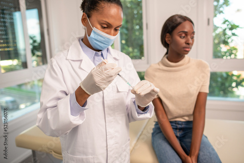 Nurse filling syringe with vaccine at doctors office. African American doctor wearing lab coat and mask preparing injection for young female patient. Vaccination and immunization concept