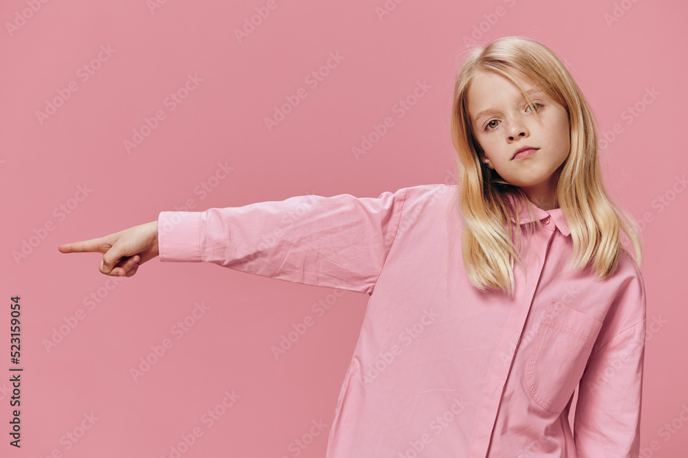 a beautiful, small, funny girl stands in a pink shirt on a pink background and points with her index finger towards the empty space on the background under an advertising mockup a beautiful, small, funny girl stands in a pink shirt on a pink background and points with her index finger towards the empty space on the background under an advertising mockup