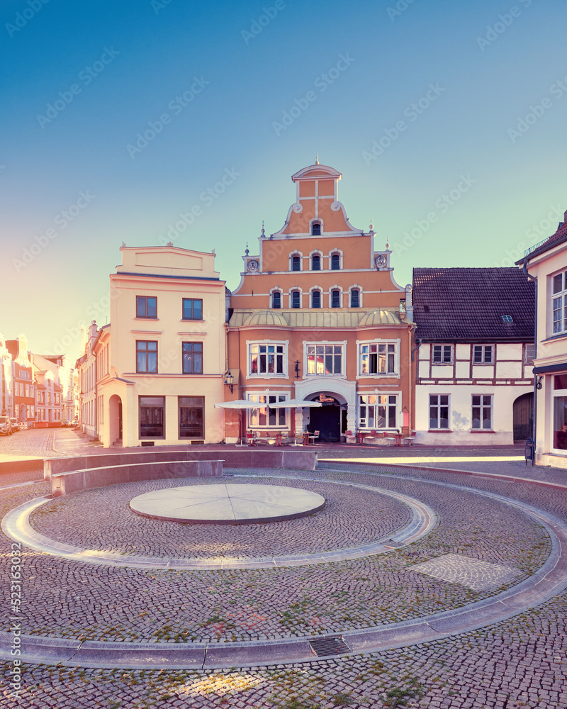 Old historic buildings in Wismar, Germany. Empty circular square early morning. Sunny day with blue sky, sun flare.