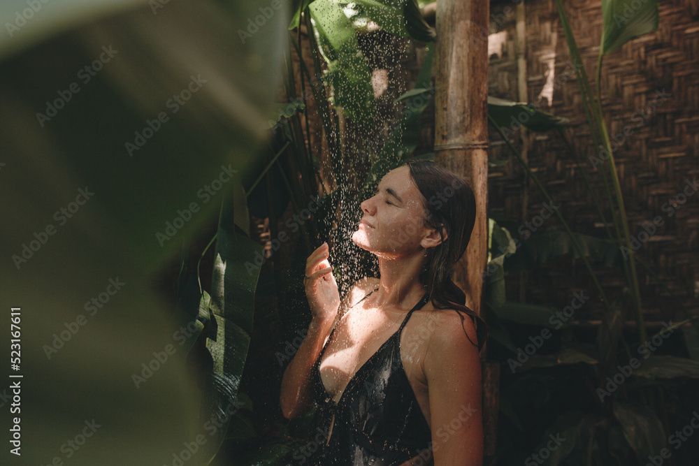 Fototapeta premium Slim woman in a black swimsuit in the shower in a tropical garden. Organic skin care. Lifestyle, holidays in Bali