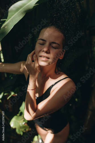 Slender young woman in a black swimsuit takes a shower outdoors in a tropical garden. Organic skin care. Lifestyle, holidays in Bali