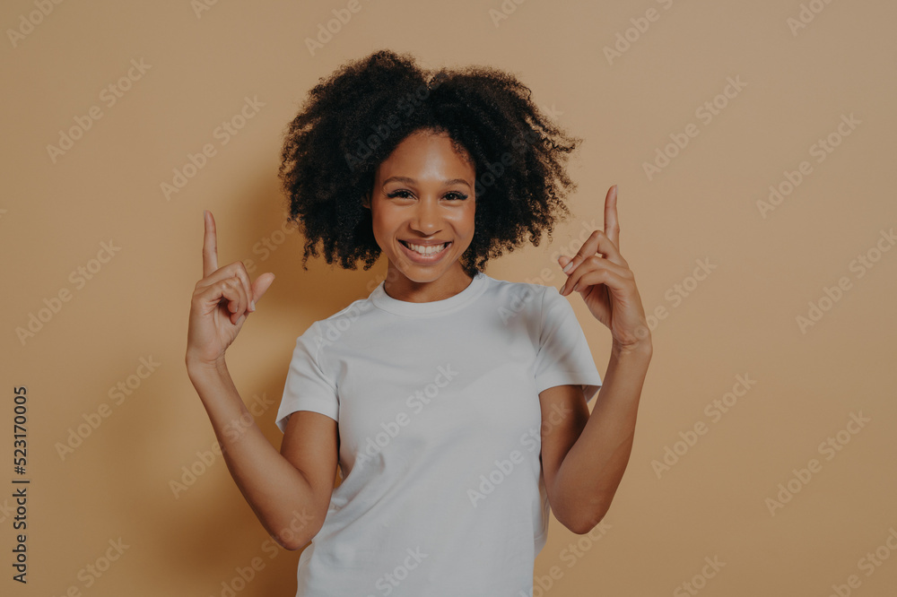 Young overjoyed dark skinned woman pointing up with forefingers, isolated on beige background Young overjoyed dark skinned woman pointing up with forefingers, isolated on beige background