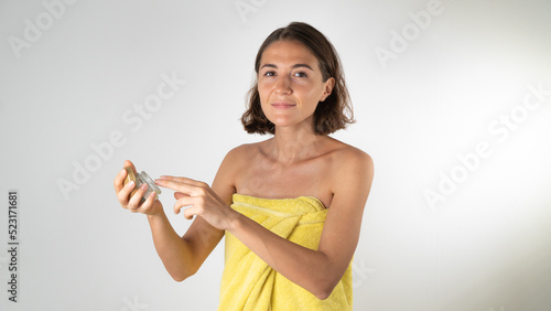 A woman in a towel applies face cream from a jar - home self-care