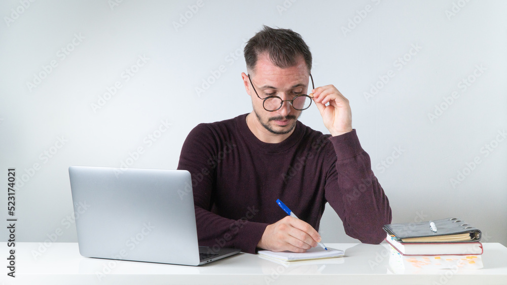 A man with glasses works at home or in the office with a laptop, makes a note in a notebook A man with glasses works at home or in the office with a laptop, makes a note in a notebook