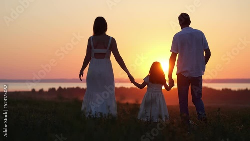 Happy parents silhouettes with excited daughter walk on evening meadow. Family looks together at bright sunset walking on lawn against rivern, slow motion photo