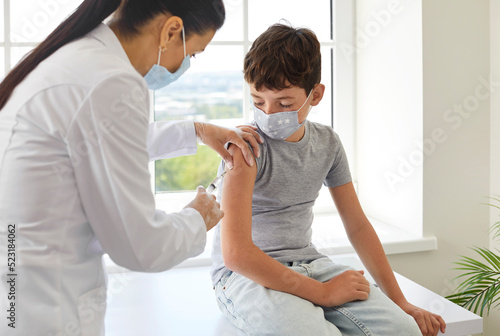 Nurse or doctor gives shoulder injection to little boy to protect him from dangerous disease. School child in medical facemask gets flu or Covid 19 vaccine at clinic during mass vaccination campaign photo