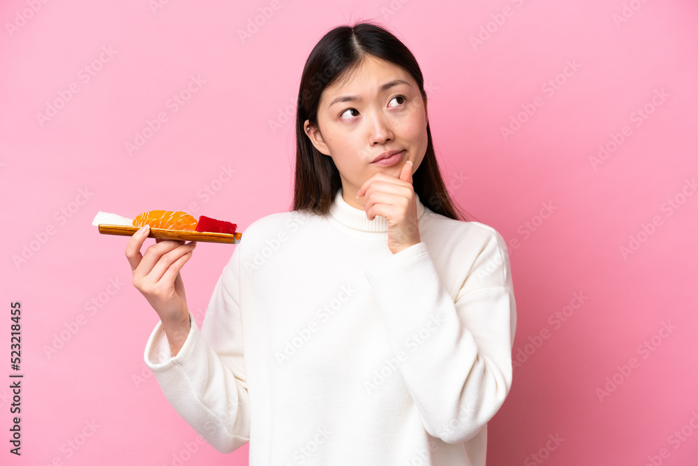 Young Chinese woman holding sashimi isolated on pink background having doubts Young Chinese woman holding sashimi isolated on pink background having doubts