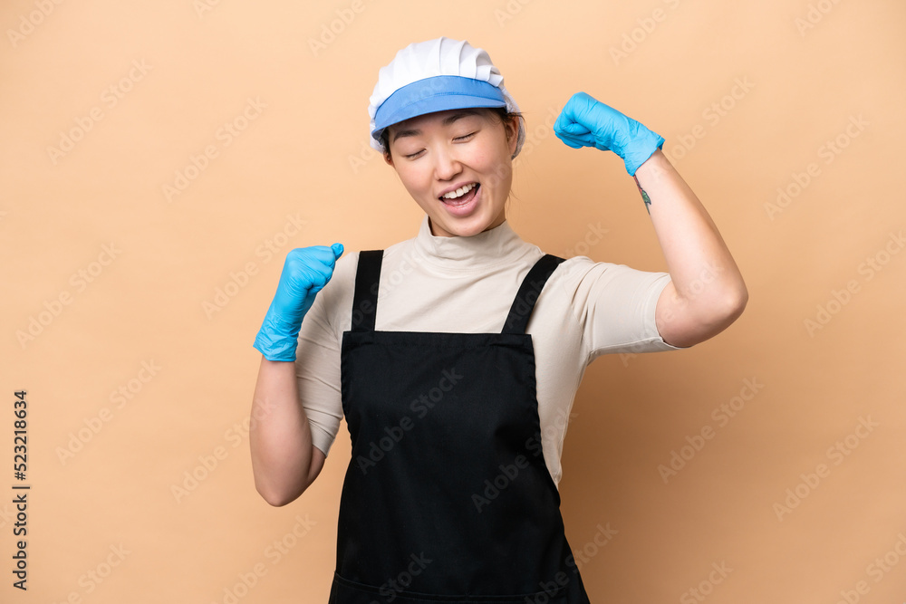 Young Chinese Fishmonger woman wearing an apron and holding a raw fish isolated on pink background celebrating a victory Young Chinese Fishmonger woman wearing an apron and holding a raw fish isolated on pink background celebrating a victory