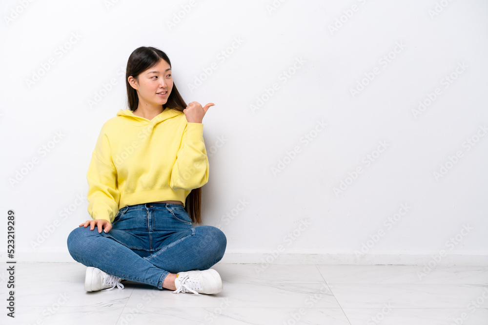 Young Chinese woman sitting on the floor isolated on white wall pointing to the side to present a product Young Chinese woman sitting on the floor isolated on white wall pointing to the side to present a product