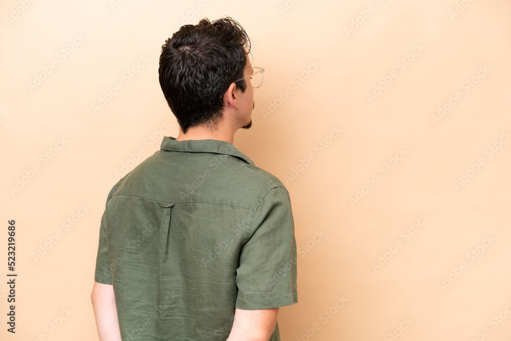 Young caucasian man isolated on beige background in back position and looking back Young caucasian man isolated on beige background in back position and looking back