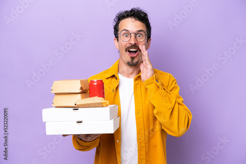 Young caucasian man holding fast food isolated on purple background with surprise and shocked facial expression