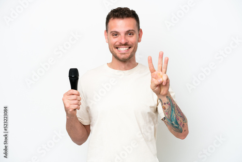 Young caucasian singer man picking up a microphone isolated on white background happy and counting three with fingers
