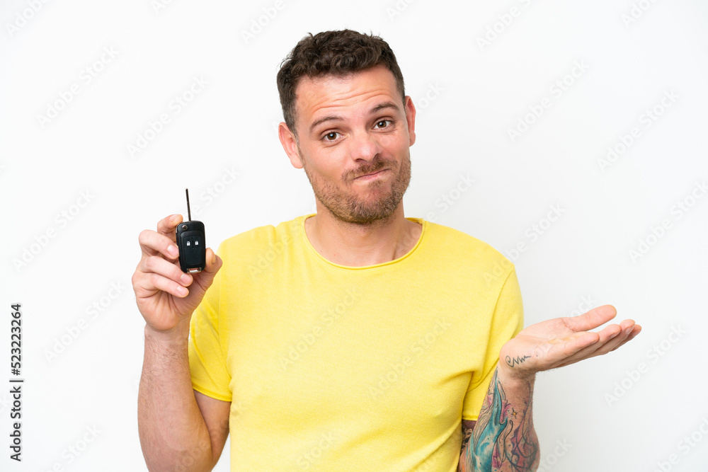 Young man holding car keys isolated on white background making doubts gesture while lifting the shoulders