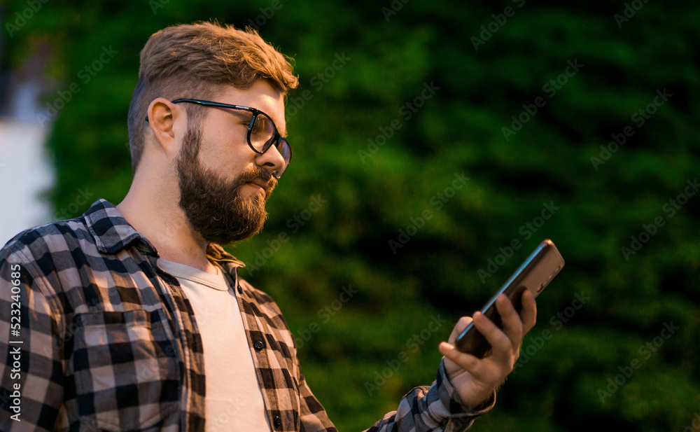 Bearded man wearing eyeglasses is holding scrolling texting in his cellphone at night street. Guy calls for taxi in an app in evening city - copy space and place for advertising Bearded man wearing eyeglasses is holding scrolling texting in his cellphone at night street. Guy calls for taxi in an app in evening city - copy space and place for advertising