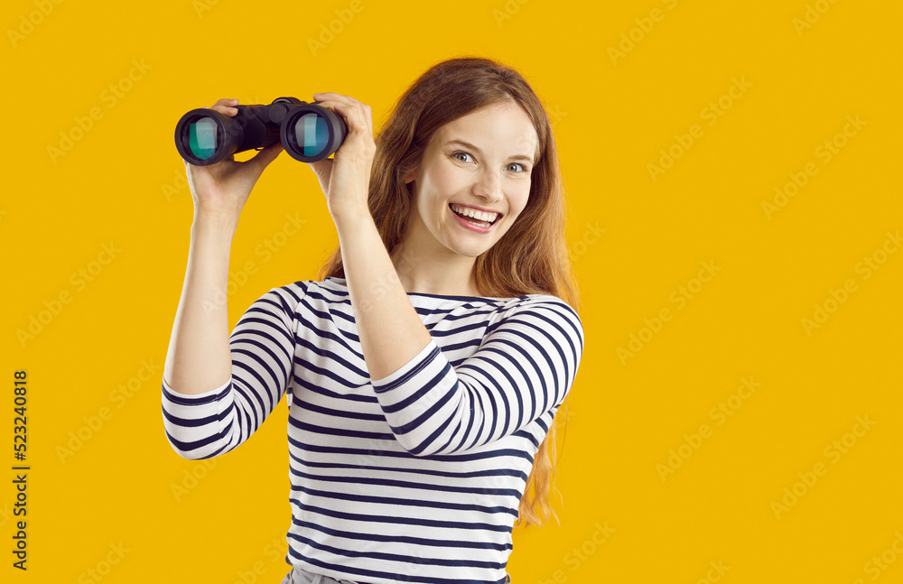Happy, smiling woman using binoculars. Casual young girl standing isolated on yellow background, holding modern binoculars and looking at camera with joyful, excited face expression. Discovery concept Happy, smiling woman using binoculars. Casual young girl standing isolated on yellow background, holding modern binoculars and looking at camera with joyful, excited face expression. Discovery concept