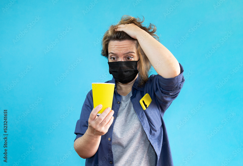 Forty year old woman in a black medical mask and shirt looks at a yellow glass of coffee on an isolated blue background. Woman portrait Forty year old woman in a black medical mask and shirt looks at a yellow glass of coffee on an isolated blue background. Woman portrait