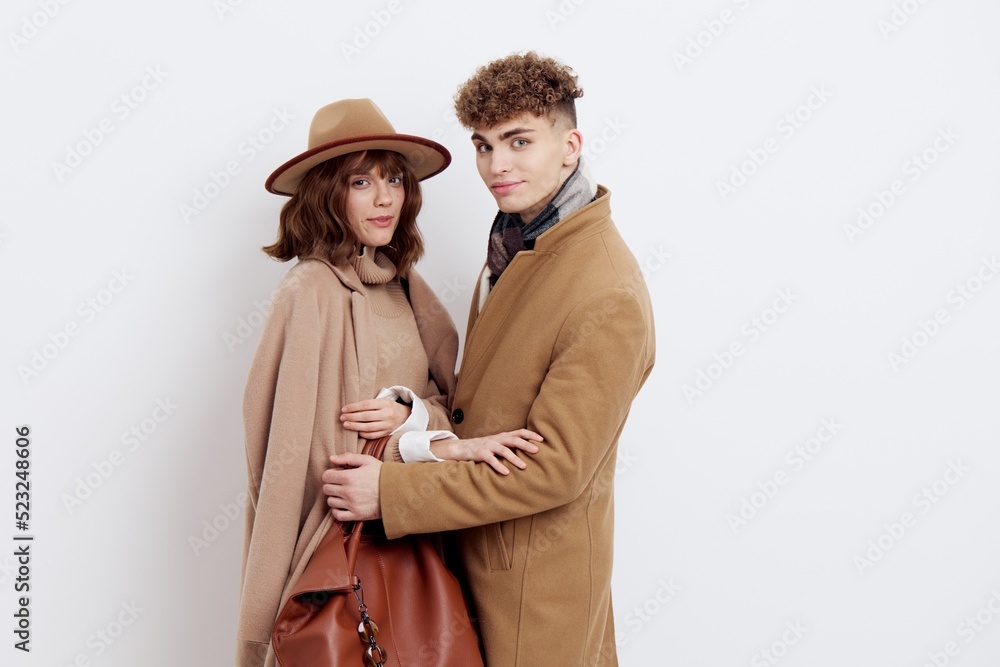 a man and a woman in an autumn coat stand on a white background and gently embrace looking at the camera, a woman has a hat on her head and she holds a bag in her hand a man and a woman in an autumn coat stand on a white background and gently embrace looking at the camera, a woman has a hat on her head and she holds a bag in her hand