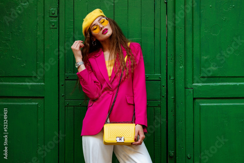 Fashionable confident woman wearing trendy outfit with yellow sunglasses, beret, wrist watch, shoulder bag, pink fuchsia color blazer, posing near green door. Copy, empty space for text