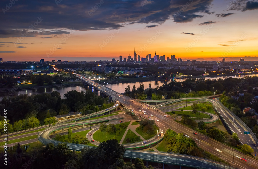 Fototapeta premium Stunning sunset skyline, aerial view Warsaw, Poland. Drone shot of city downtown business center skyscrapers in background. Highway bridge over river and driving cars, amazing cloudscape evening dusk