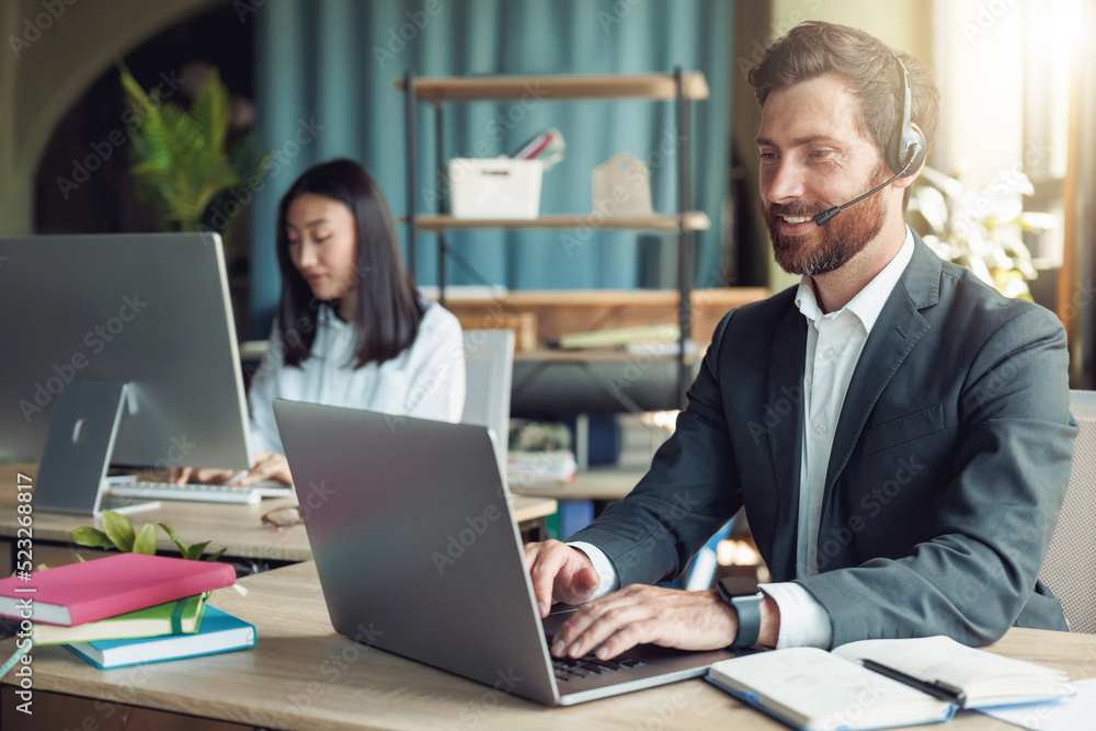 Fototapeta premium Male operator using headset and computer while talking with customers online on colleague background