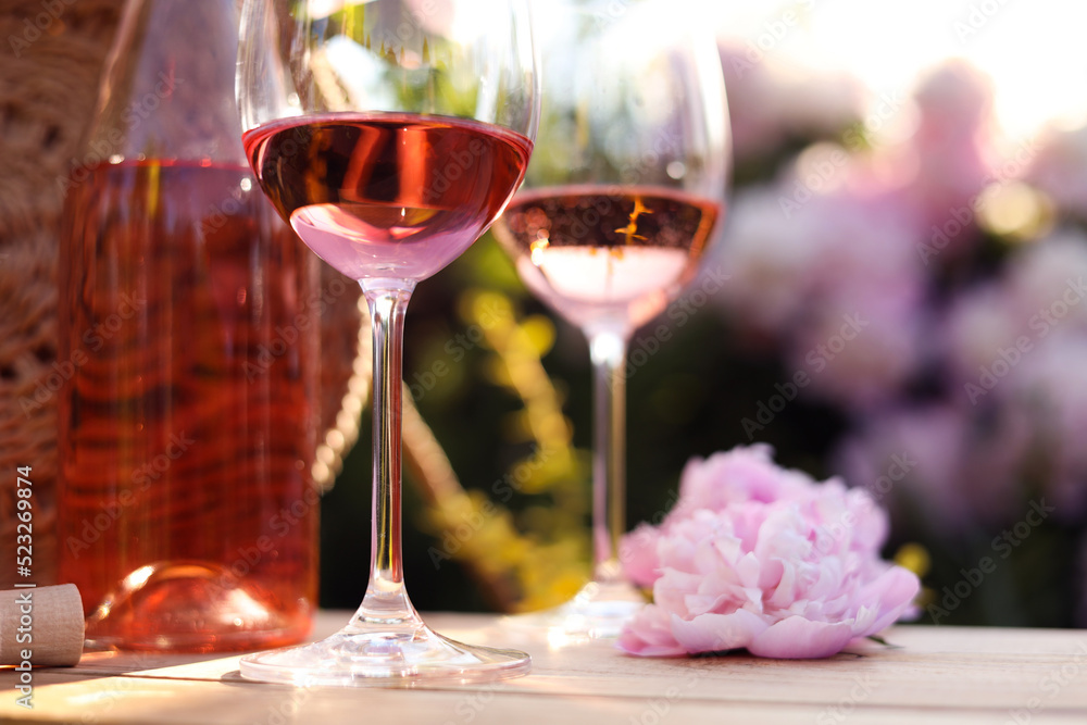 Bottle and glasses of rose wine near beautiful peony flower on wooden table in garden, closeup