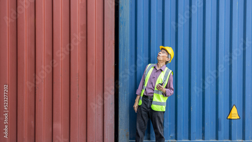 Elderly supervisor stands next to a blue container using walkie talkie to advise his coworkers in a shipyard or logistic station. Senior advisor communicates with colleagues in a storage place.