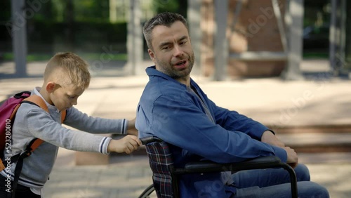 Side view of the son went for a walk with his disabled father and pushes his wheelchair and father telling interesting stories for his son. Family and disabled people concept photo