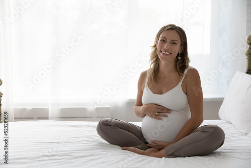 Beautiful young 35s pregnant woman in comfort home-wear touch big belly in late pregnancy smile look at camera sit on bed in light bedroom at home. Happy expectant mother portrait, maternity concept.