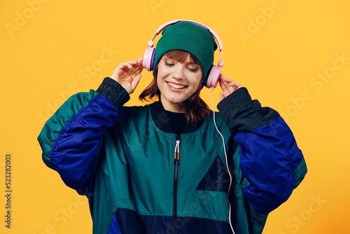 a happy, joyful woman in a green jacket and hat enjoys music standing in pink headphones on a yellow background holding them with her hands and looking away with a pleasant smile on her face