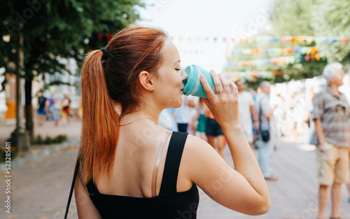 Young woman outdoors summer festival holiday lifestyle. Rear view of cute woman drinking coffee while standing at an event, crowd of people walking along decorated street in bokeh