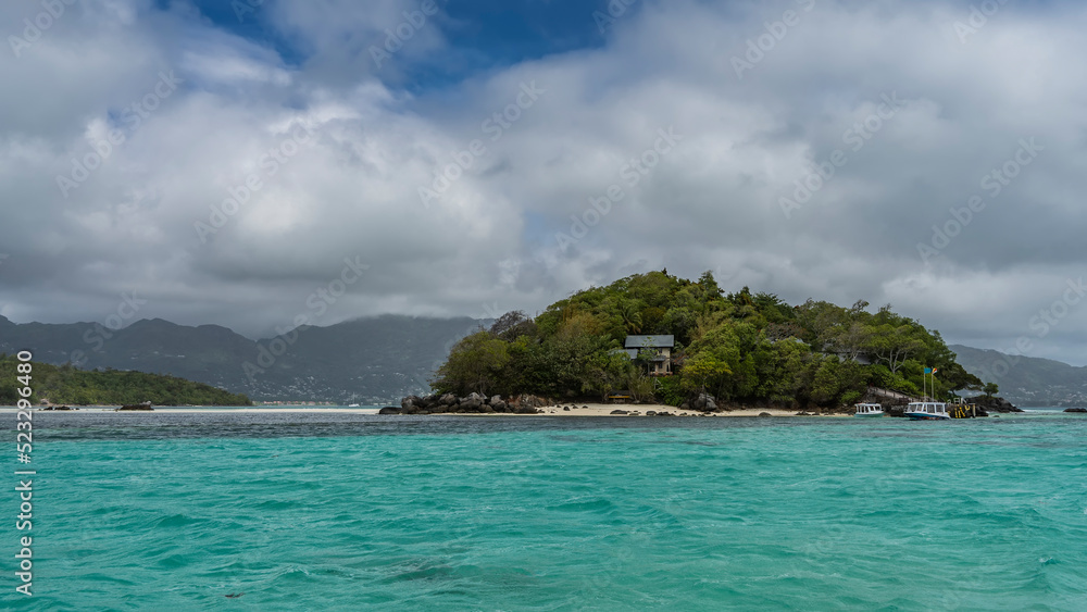 The tropical island is overgrown with lush vegetation. Villas can be seen through the foliage. Boats are moored at the sandy beach. Turquoise ocean and blue sky with clouds. Seychelles