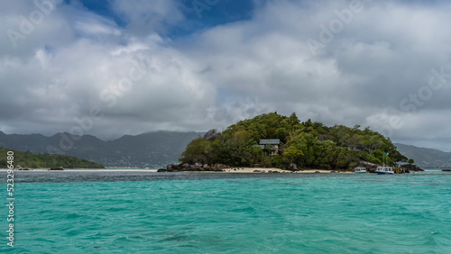 The tropical island is overgrown with lush vegetation. Villas can be seen through the foliage. Boats are moored at the sandy beach. Turquoise ocean and blue sky with clouds. Seychelles