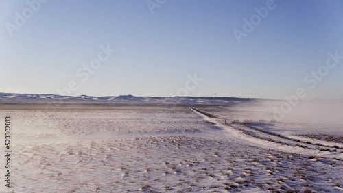 Aerial drone view of a snow-covered dirt road cutting through a remote winter landscape in Mammoth Lakes, California. Vast open terrain, distant mountains, and a clear blue sky create a minimalist sce