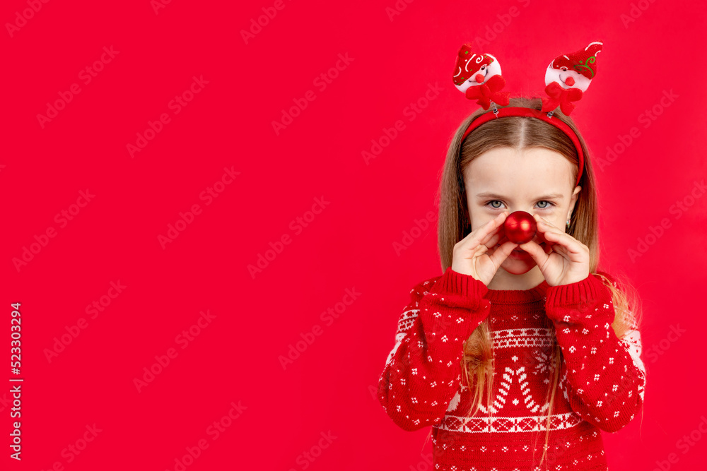 a child girl in a sweater with a ball on her nose on a red monochrome isolated background rejoices and smiles, the concept of new year and Christmas, space for text