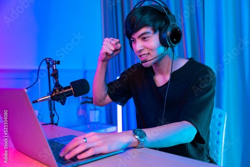 Playing video games on smartphone. Young asian handsome man sitting on chair holding cellphone in his hand. Exited streamer wearing headset in neon room .Esport streaming game online.