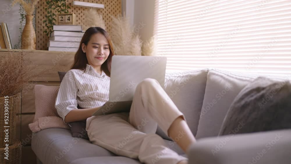 Smiling Asian freelance business women casual wear sitting on sofa using laptop looking at screen typing message,working call video conference in living room.Happy young girl relax at home in summer Smiling Asian freelance business women casual wear sitting on sofa using laptop looking at screen typing message,working call video conference in living room.Happy young girl relax at home in summer
