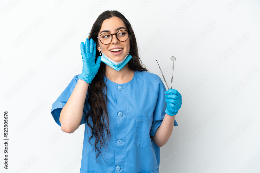 Young woman dentist holding tools isolated on white background listening to something by putting hand on the ear